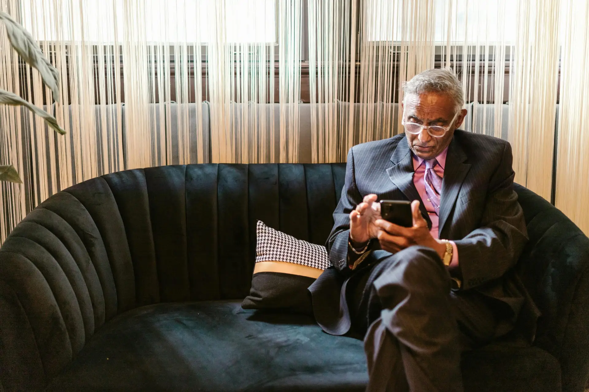 Elderly man in a suit engaging with a smartphone while seated indoors.