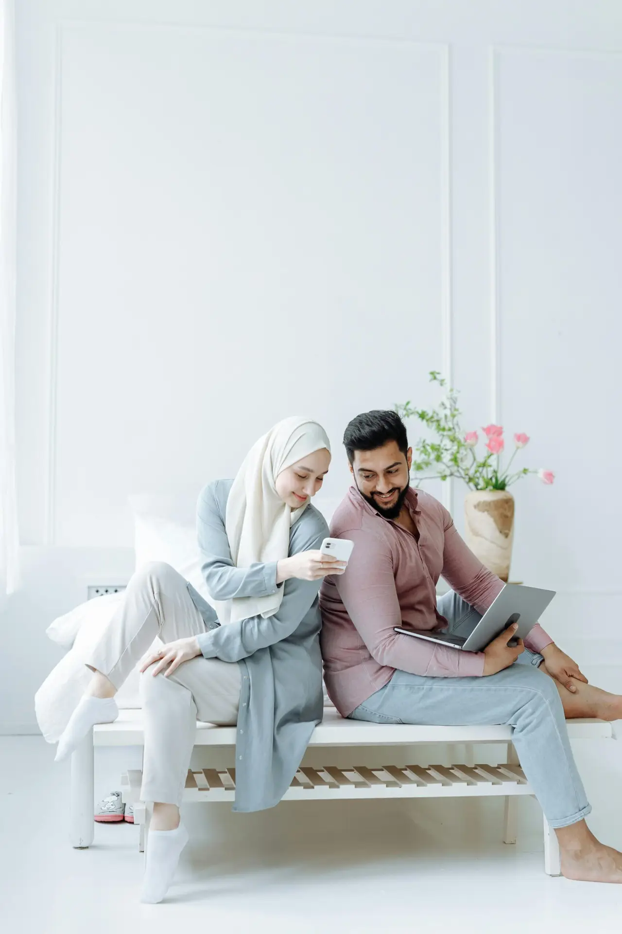 Young Muslim couple sitting back to back, using devices in a bright room.